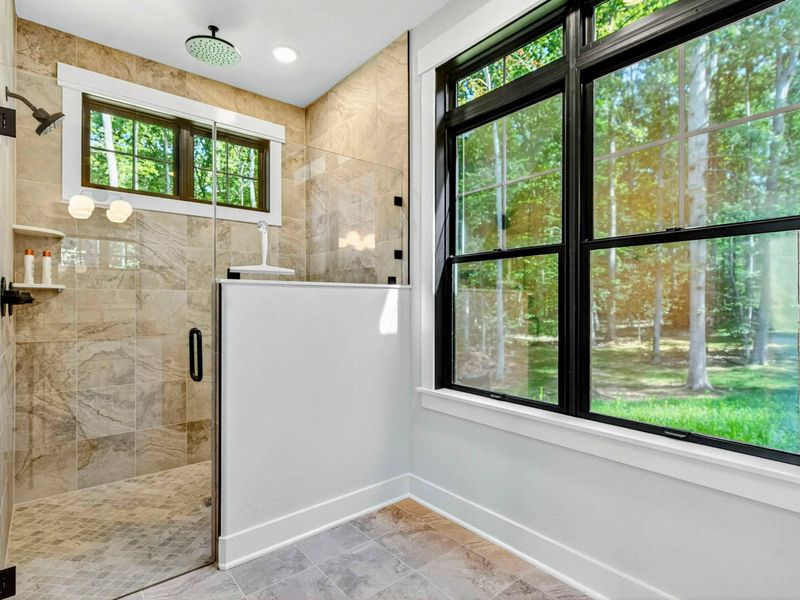 Primary Bathroom - Walk-in shower with rain head, warm marble-look tile, and a large window to the trees.