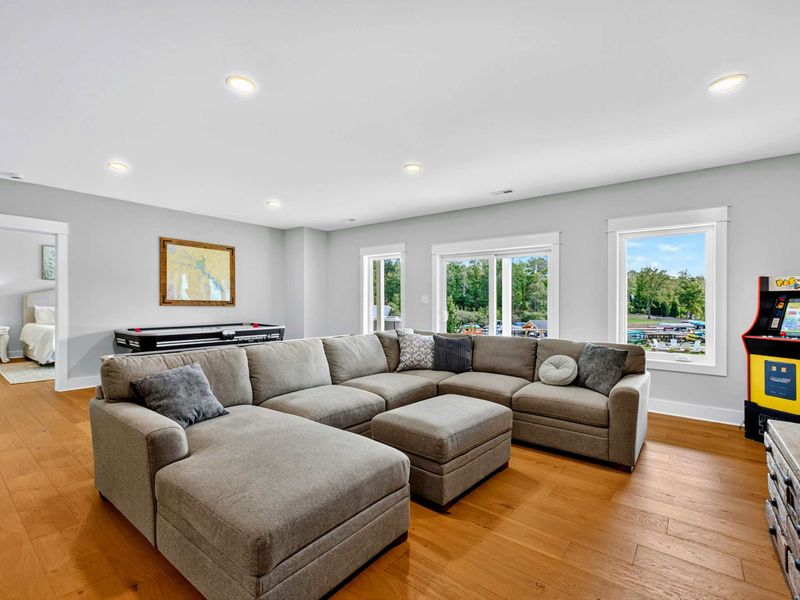 Rec Room Seating - A deep sectional with lake views through the windows, a Pac-Man arcade cabinet, shuffleboard table, and a doorway to the lower-level bedrooms.