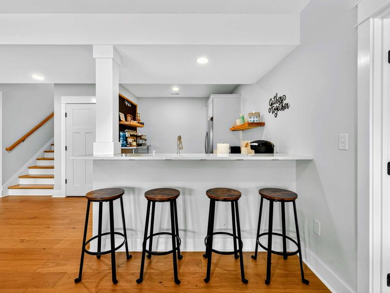 Second Kitchen Bar - Four bar stools line the lower-level kitchen counter, with a coffee bar, "Gather Together" sign, and the staircase to the main level.