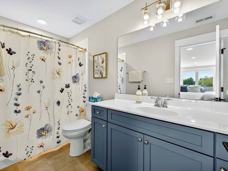 Upper Ensuite - A navy blue vanity with quartz countertop, gold sconce lighting, floral shower curtain, and the full size window seat/bed reflected in the mirror.