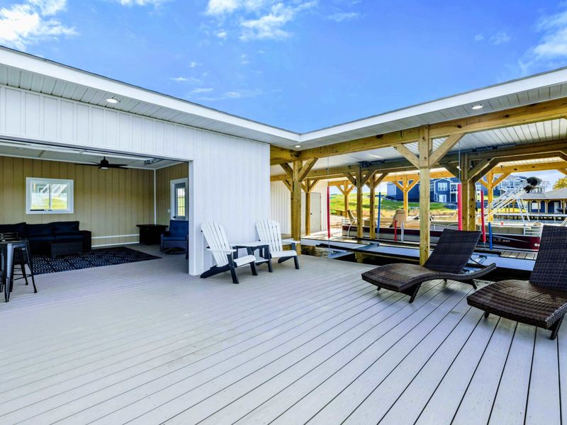 A spacious lakeside deck with lounge chairs, Adirondack seating, and a shaded boat slip area, seamlessly connected to a cozy indoor lounge