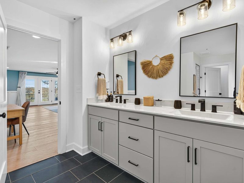 Primary Ensuite Vanity - A double vanity with gray cabinets, quartz tops, black fixtures, and a view back into the primary suite.