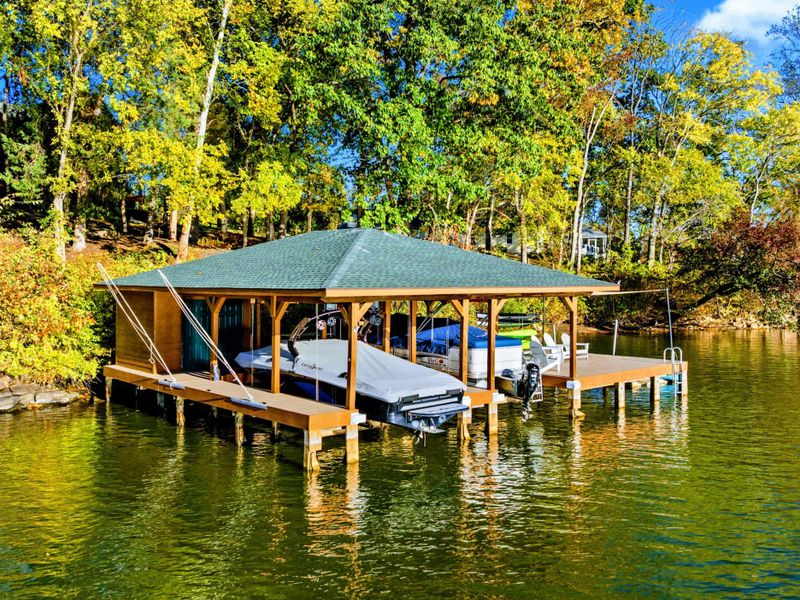 Aerial - Covered dock and boathouse with a green metal roof viewed from the water, framed by fall foliage along the shoreline.