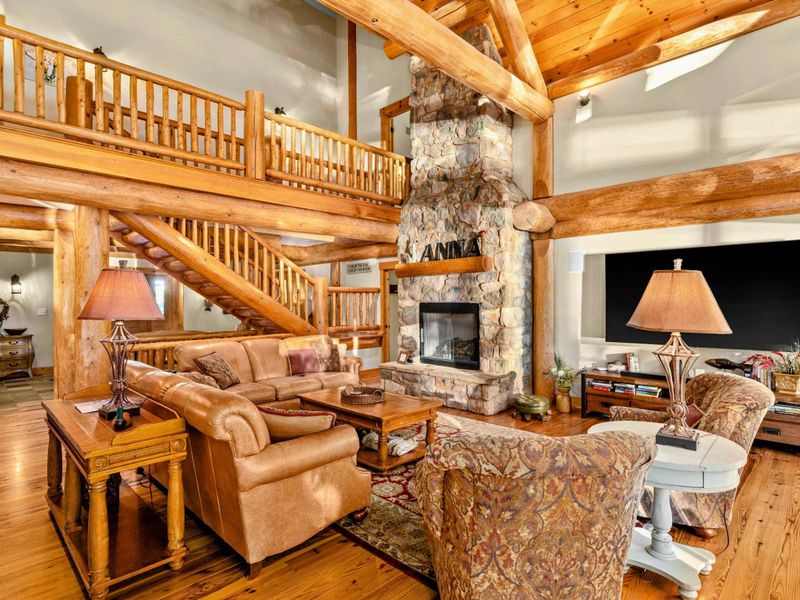 Great Room - Soaring two-story space with a stone fireplace, exposed log beams, loft railing above, and leather seating on hardwood floors.