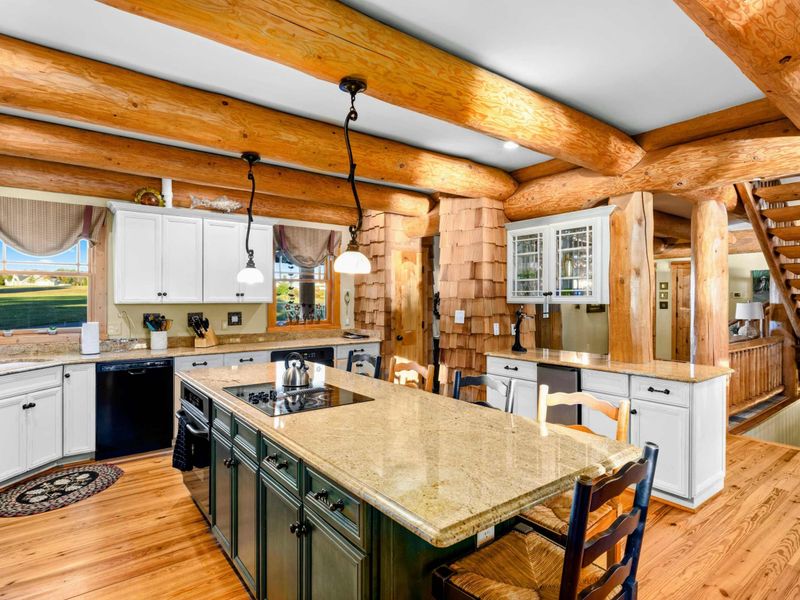 Kitchen - Large island with a cooktop, pendant lights, and bar seating under massive log beam ceilings with white and green cabinetry.