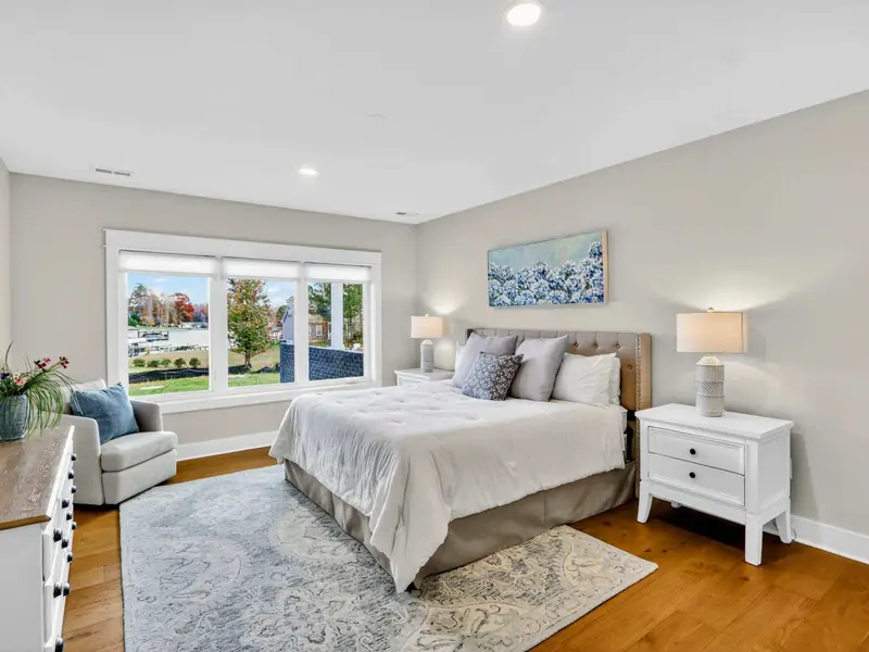 Lower Level King Suite - A king bed with a tufted gray headboard, blue hydrangea artwork, accent chair, and windows overlooking the lawn and lake.