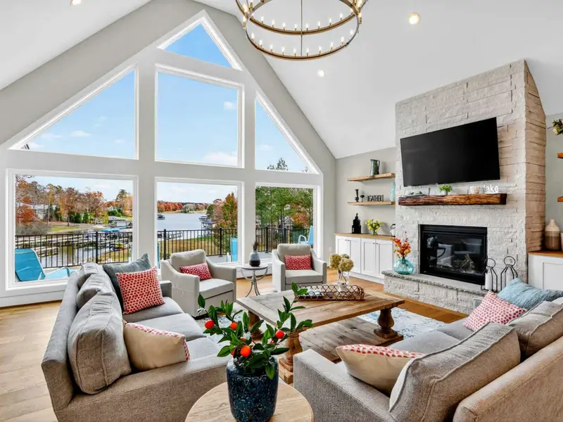 Great Room - Vaulted ceilings and a wall of lake-facing windows fill the living room with natural light, anchored by a white brick fireplace and rustic mantel.