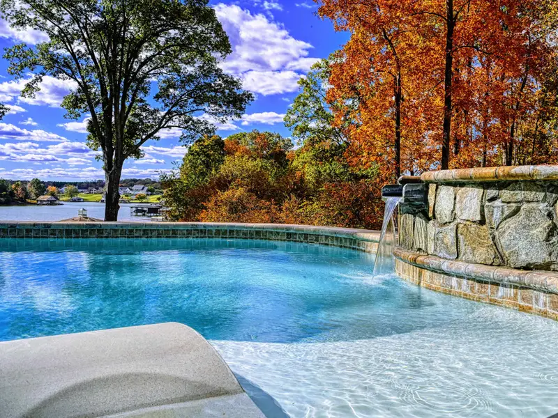 Resort-style heated pool with a built-in spa shelf and baja loungers in the water, framed by full lake views.