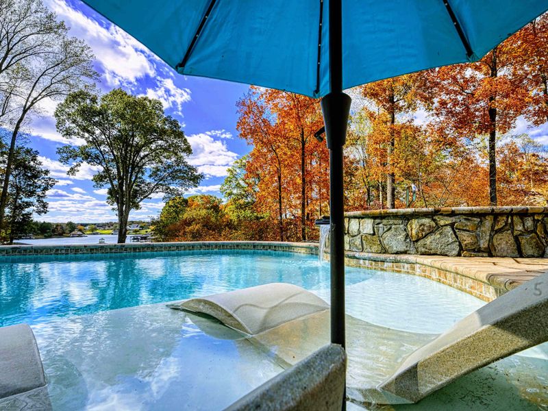 Resort-style heated pool with a built-in spa shelf and baja loungers in the water, framed by full lake views.