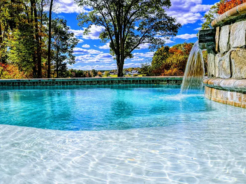 Resort-style heated pool with a built-in spa shelf and baja loungers in the water, framed by full lake views.