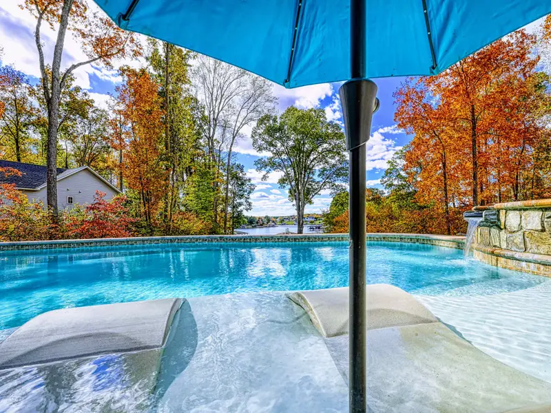 Resort-style heated pool with a built-in spa shelf and baja loungers in the water, framed by full lake views.