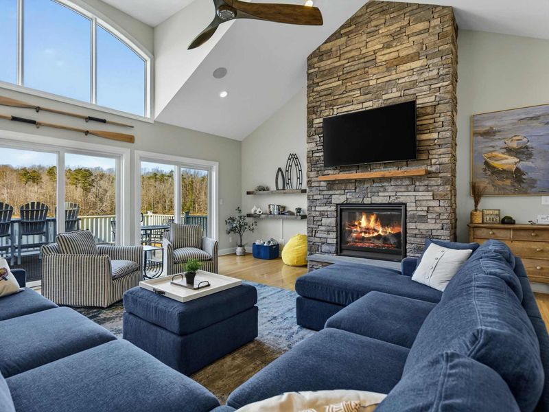 Great Room - Vaulted ceilings, a floor-to-ceiling stone fireplace, and navy sectionals beneath a wall of windows opening to the deck and lake.