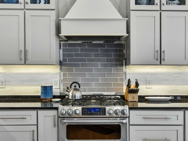 Kitchen - Close-up of the gas range and custom hood surrounded by gray tile and gray cabinetry.