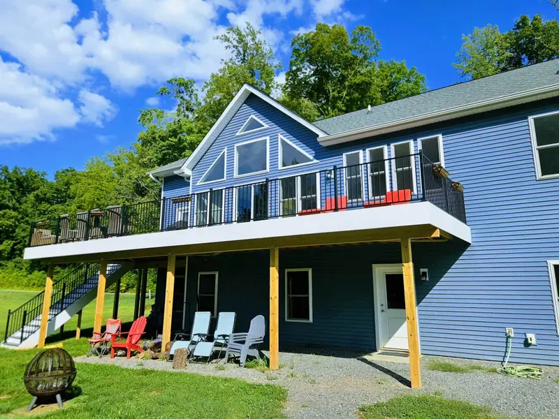 Exterior - Blue-sided lakefront home featuring A-frame peak windows, an upper deck with black railings, and a lower covered patio with Adirondack seating.