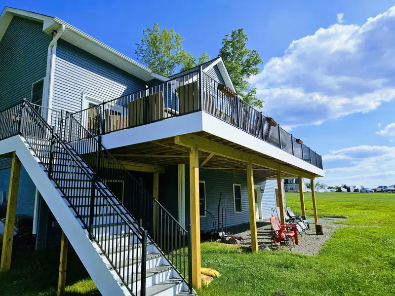 Exterior - Rear view showcasing the upper deck with black railings, lower covered patio with red Adirondack chairs, and stairway connecting both levels.