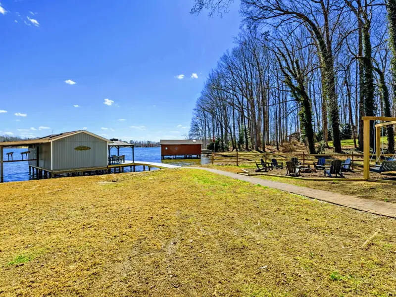 Lakeside Grounds - The full outdoor setup comes together: boathouse, dock, fire pit, bench swings, and flat yard stretching back to the house.