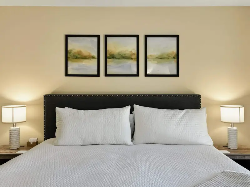 Primary Suite Detail - Three watercolor landscape prints hang above the upholstered headboard, framed by matching ceramic lamps on rustic nightstands.