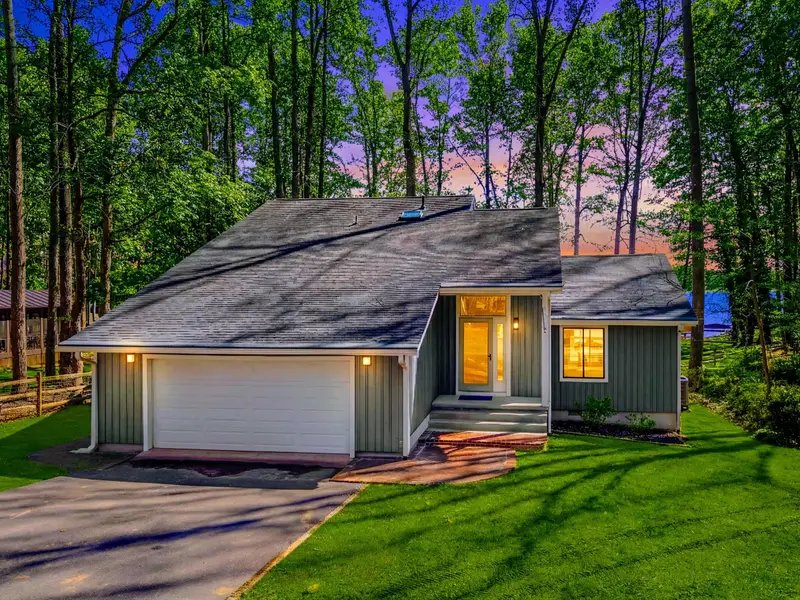 Evening Exterior - Warm light glows from inside as dusk settles over the lawn and wooded surroundings.