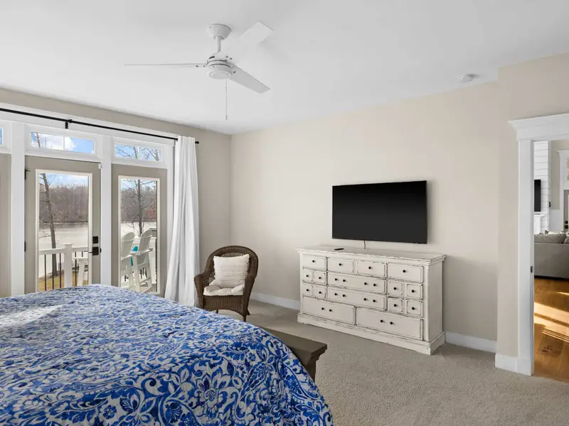 Primary Suite - King bed, TV on white dresser, ceiling fan, and a wicker chair, with French doors bringing the lake inside.