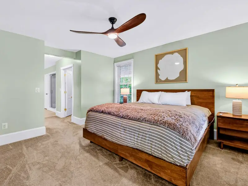 Primary Suite - King bed framed by sage-green walls, ceiling fan, and quiet morning light through large windows.