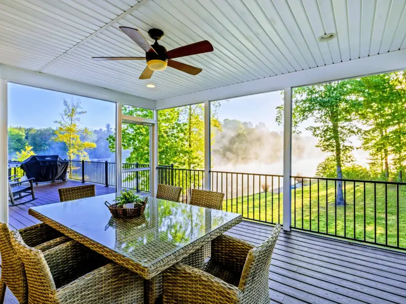 A cozy screened-in porch with a wicker dining table for six, ceiling fan, and morning lake views, with the grilling deck just beyond.