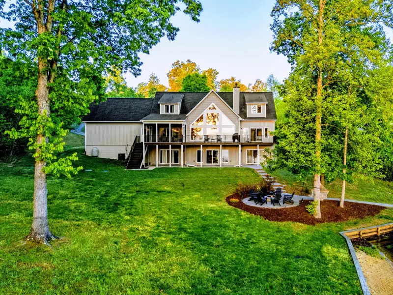 An aerial view of the back of the home showcasing the deck, screened porch, lower patio with fire pit, sloping lawn, and lush wooded surroundings.