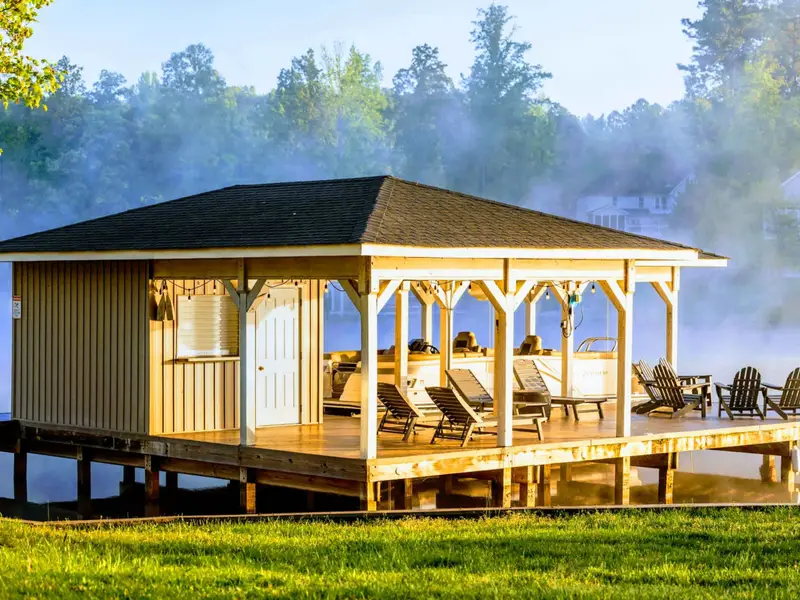 A picturesque covered boathouse and dock featuring lounge chairs, a pontoon boat slip, and Adirondack seating, framed by mist rising off the lake at sunrise.