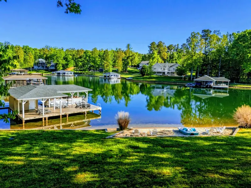 A panoramic view of the cove from the backyard featuring the private covered dock, sandy beach with Adirondack seating, and the quiet community shoreline across the water.
