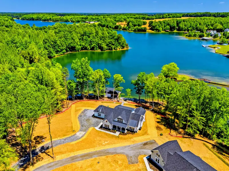 Aerial - Overhead look at the gray-roofed home on its cleared lot, framed by dense green forest and a glassy finger of the lake just beyond the trees.