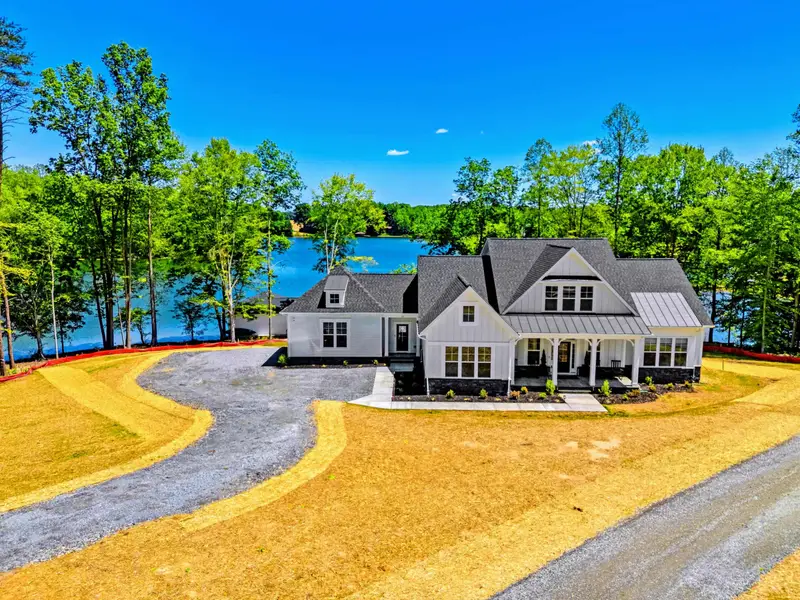 Exterior - Ground-level view of the white board-and-batten farmhouse with its covered porch and gabled roofline, the lake shimmering through tall pines behind.