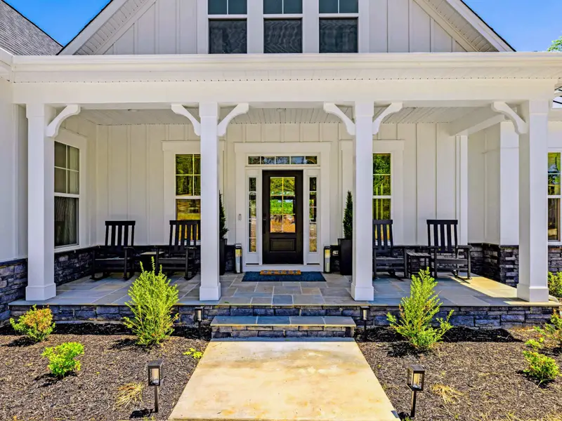 Front Porch - Inviting covered entrance framed by white columns and stone bases, with black rocking chairs, potted topiaries, and a bluestone walkway leading to the glass front door.