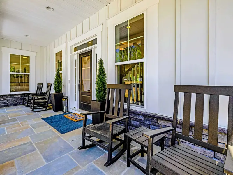 Front Porch - Sunlit flagstone porch with paired black rocking chairs, potted evergreens, and a blue runner welcoming guests to the glass-paneled door.