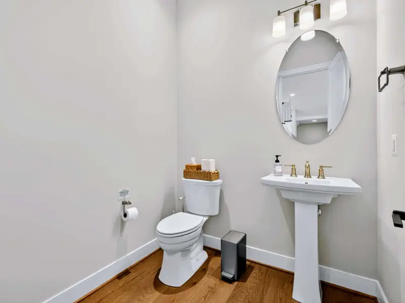 Half Bath - A pedestal sink with brushed gold faucet sits beneath an oval mirror and triple sconce, finished with warm hardwood floors.