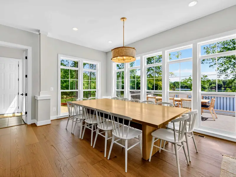 Dining Area - Light oak farmhouse table with white spindle chairs sits before a wall of windows revealing the deck and shimmering lake water.
