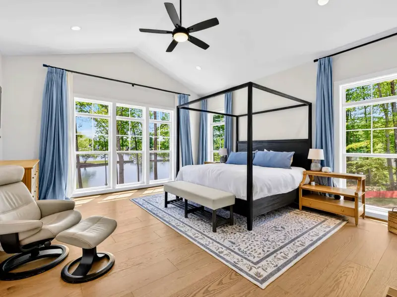 Primary Suite - A black four-poster king anchors the vaulted retreat, framed by floor-to-ceiling lake windows, soft blue drapes, and a leather recliner by the water view.