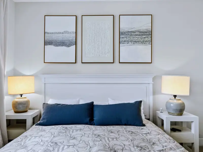 Bedroom 3 - Headboard view of the king bed with a navy bolster, twin ceramic lamps on white side tables, and three woven-texture prints above.