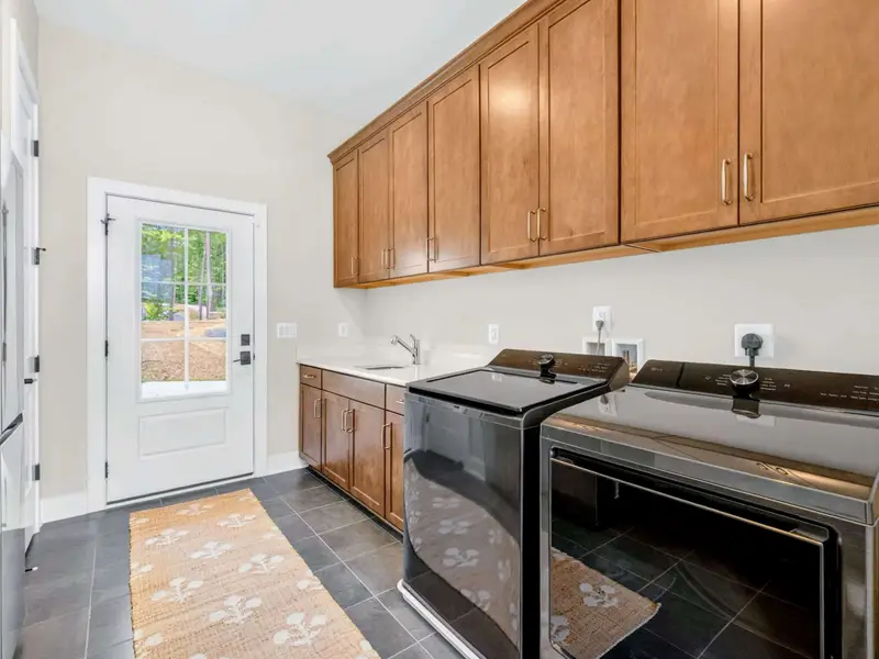 Laundry - Full-size top-load washer and dryer beside a counter with sink and warm wood cabinetry, plus an exterior door to the yard.