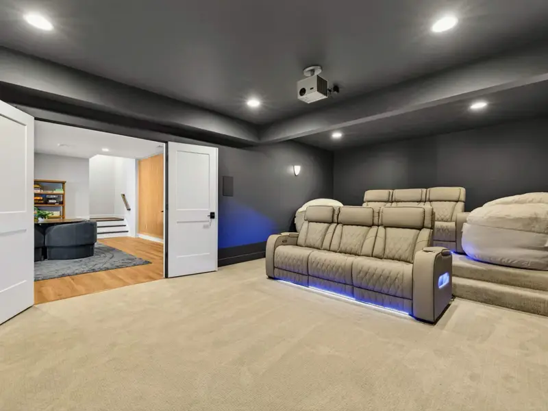 Movie Theater - Tiered home cinema with plush cream power recliners glowing under blue LED lighting, oversized bean bags, and a ceiling projector ready for showtime.