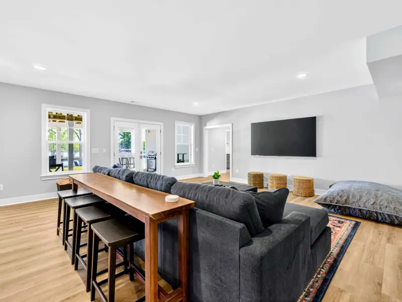 Game Room - Long live-edge bar table with stools behind a charcoal sectional, wall-mounted TV, woven poufs, and floor cushions beside lakeview French doors.