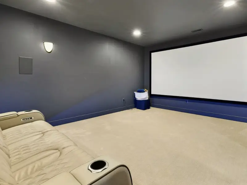 Movie Theater - Wide projection screen on a deep gray wall faces a cream leather recliner with cup holders across the plush carpeted floor.