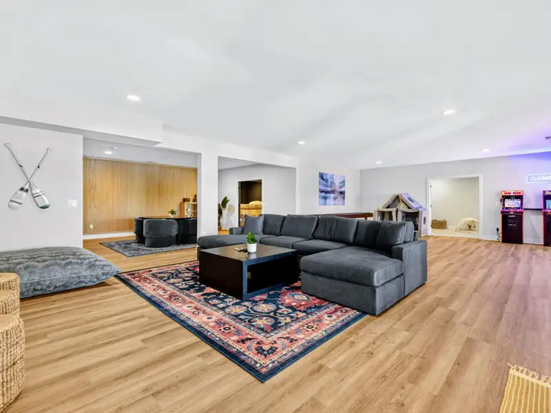 Game Room - Expansive open space with crossed-paddle wall art, floor cushions, a sectional on a floral rug, and arcade cabinets beneath the GAME ON neon.