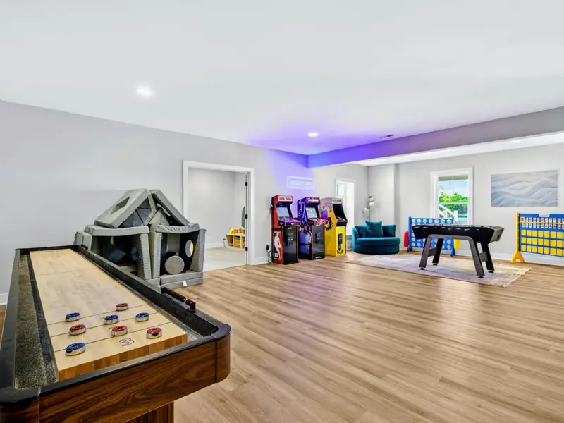 Game Room - Detail of the wood shuffleboard surface scored with pucks and bead counter, with NBA arcade games standing in the sunlit play zone beyond.