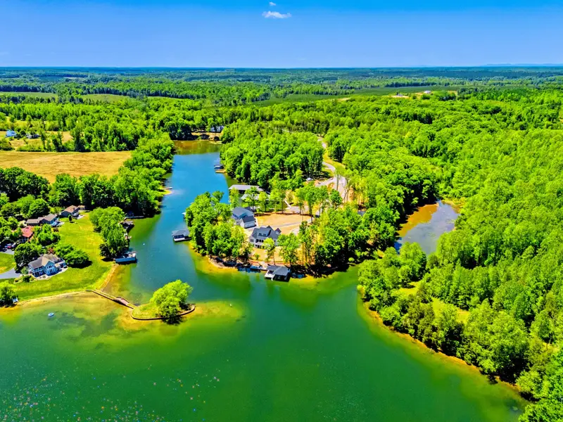 Aerial - Expansive high view of the home set among coves and forest, with open water, distant farmland, and rolling tree canopy reaching the horizon.