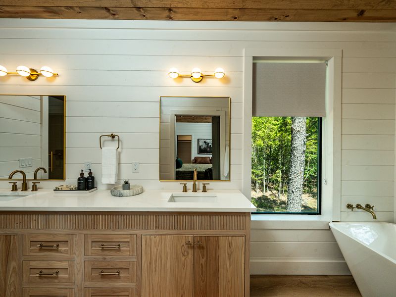A spacious primary bathroom features a double vanity, gold fixtures, a soaking tub, and beautiful views of the surrounding woods.