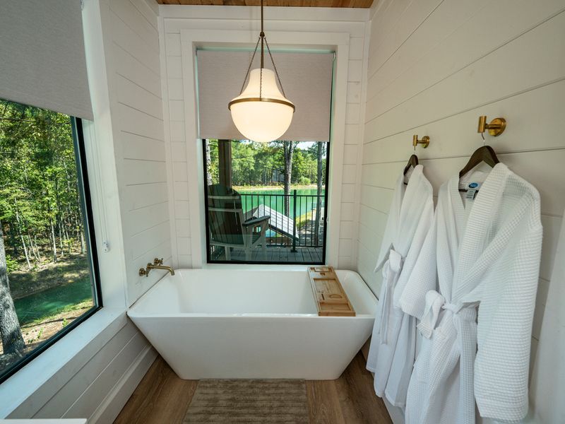 A freestanding soaking tub sits beside large windows overlooking the peaceful lake and wooded surroundings.