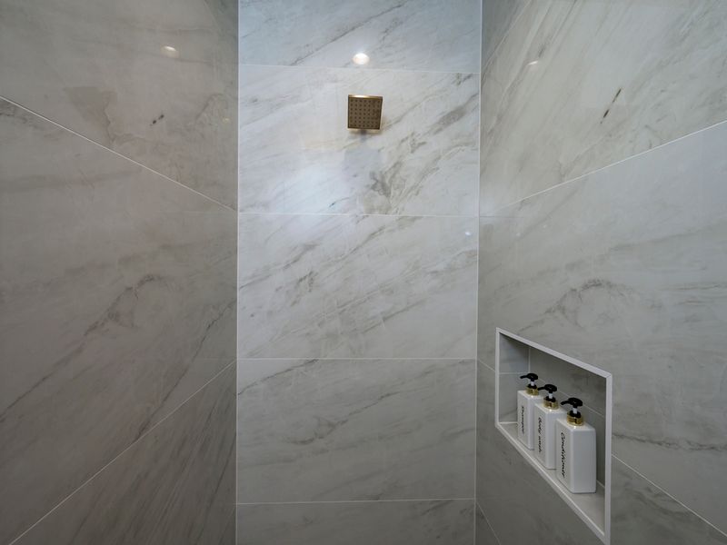A spacious walk-in shower is finished with elegant marble-look tile, a rainfall showerhead, and convenient built-in shelving.