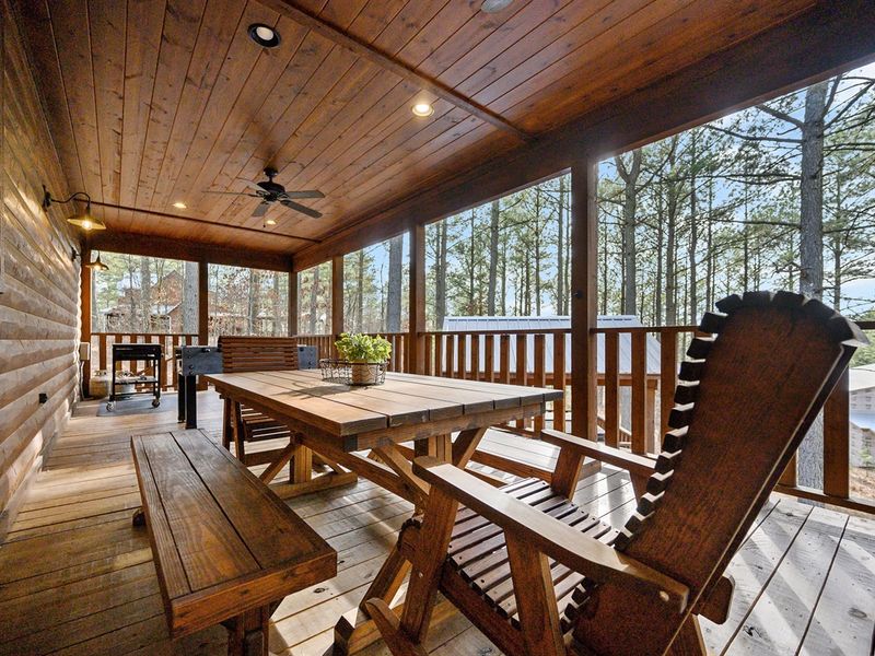 Cozy wooden porch featuring a large dining table, comfortable seating, and surrounded by scenic trees
