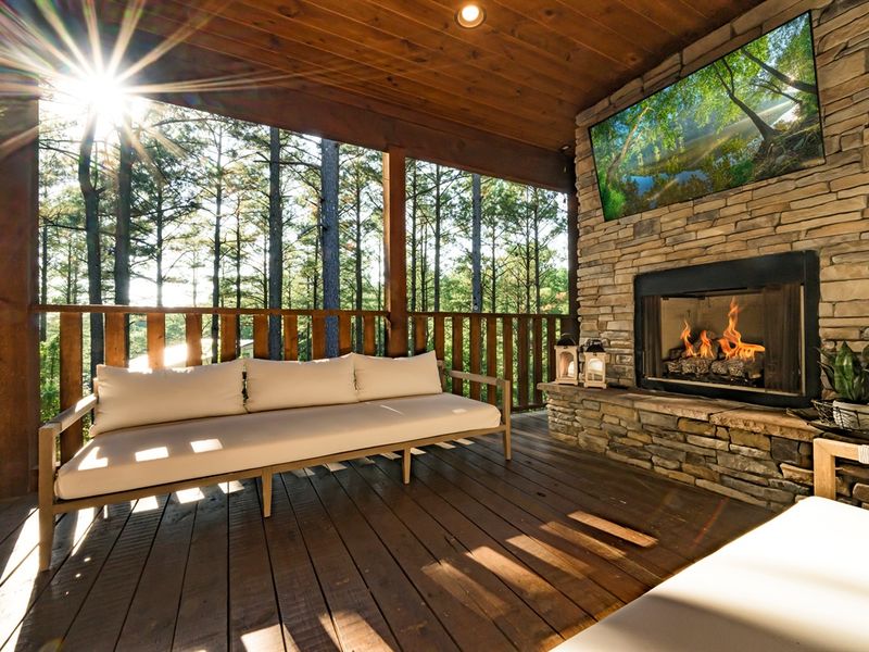 Cozy outdoor seating area featuring a stone fireplace and a mounted TV, surrounded by tall