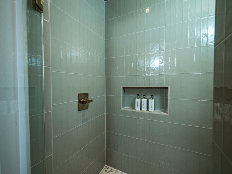 Glass-enclosed walk-in shower featuring soft green tile, a pebble floor, and built-in shelving.