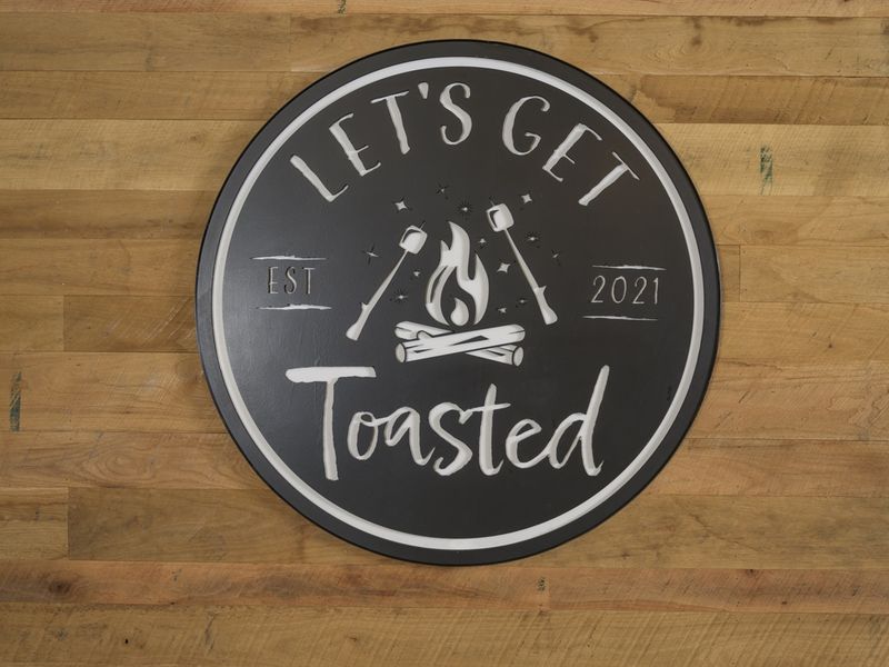 Round black sign with the phrase "LET'S GET Toasted" and a campfire graphic, established